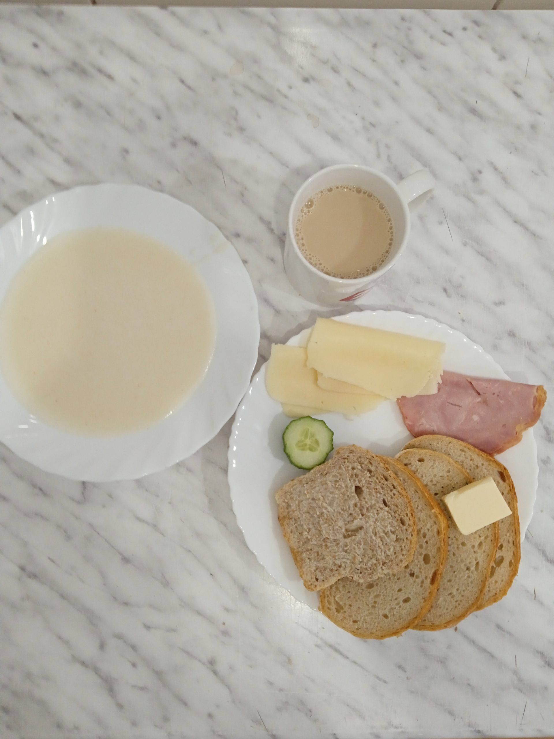 zdjęcie przedstawia śniadanie złożone z zupy mlecznej, trzech kromek chleba, masła, dwóch plastrów sera żółtego i szynki, oraz ogórka zielonego. Posiłki ułożone na dwóch białych talerzach. Powyżej talezy kubek z kawą zbożową. 