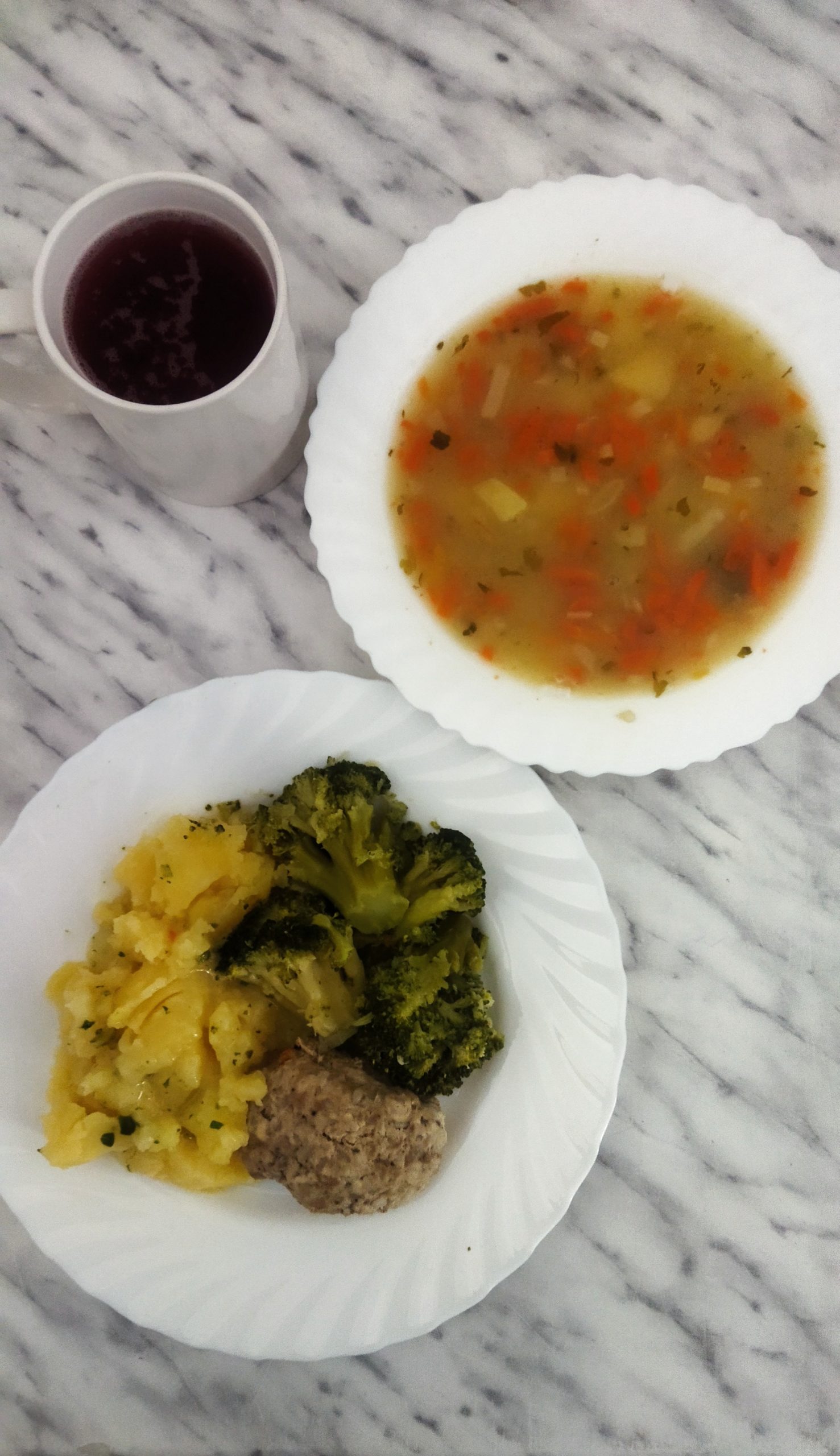 na zdjęciu obiad złożony z dwóch dań. Na talerzu głębokim zupa jarzynowa, na talerzu płaskim ziemniaki polane sosem mięsnym, pulpet i brokułu. Powyżej talerzy kubek z kompotem