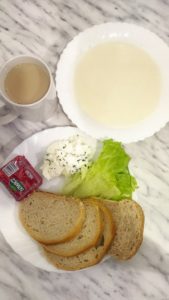 na zdjęciu śniadanie podane na dwóch talerzach. W glębokim zupa mleczna, na płaskim cztery kromki chleba, sałata, twarożek ze szczypiorkiem i drzem. Do śniadania kubek z kawą zbożową