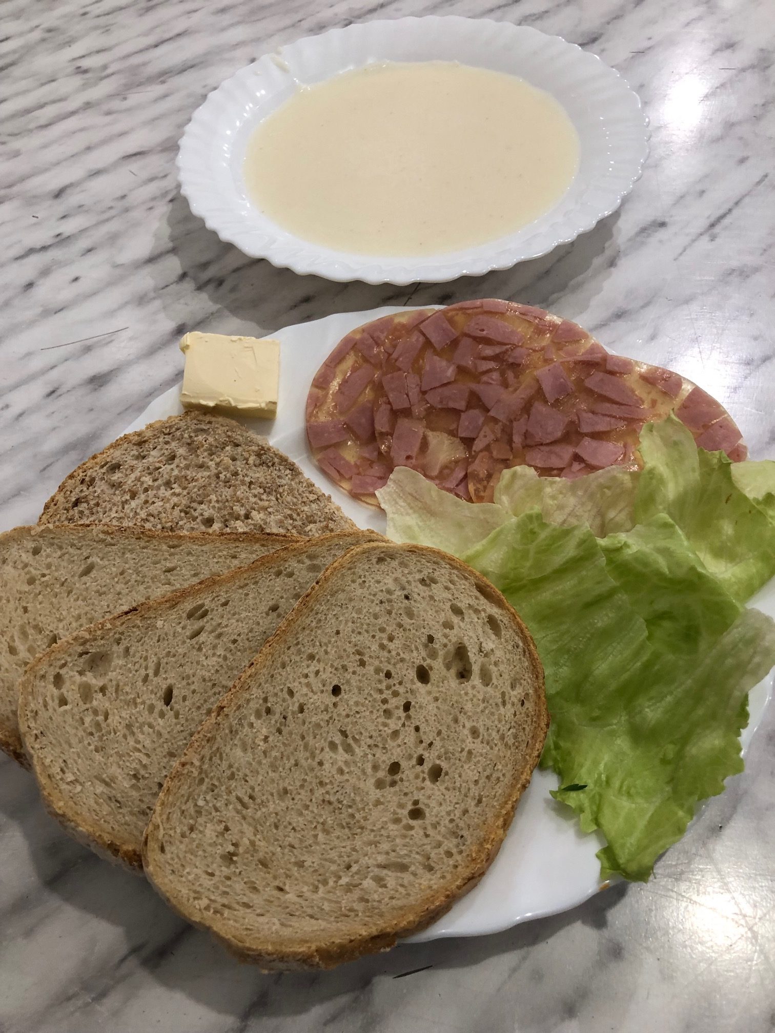 zdjęcie przedstawia śniadanie zlozone z czterech skibek chleba, dwóch plastrów szynki, masła i liści sałaty. na drugim talerzu powyżej zupa mleczna