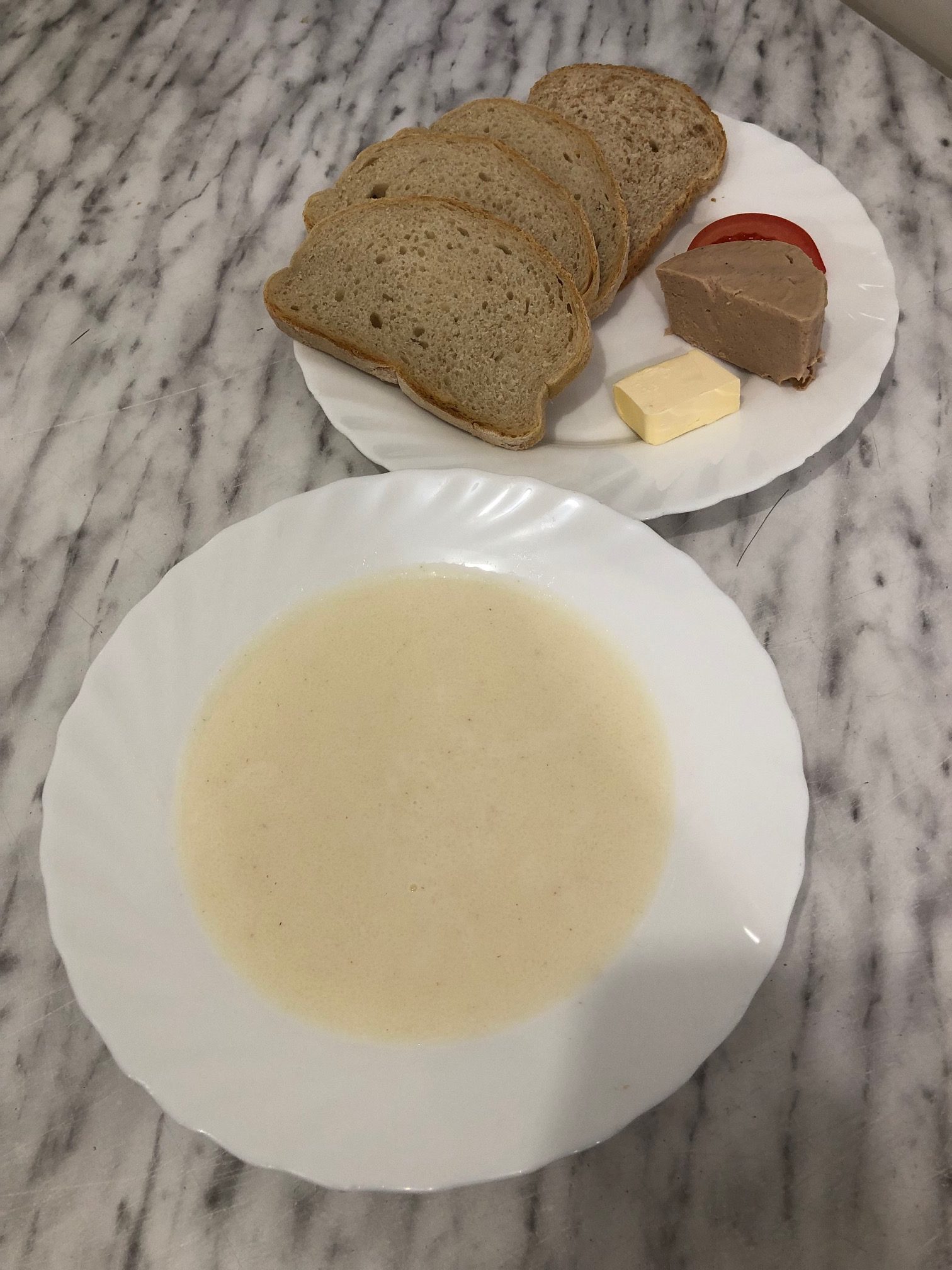 zdjęcie przedstawia śniadanie złożone z czterech skibek chleba, pasztetu, pomidora i masła. Na drugim talerzu zupa mleczna