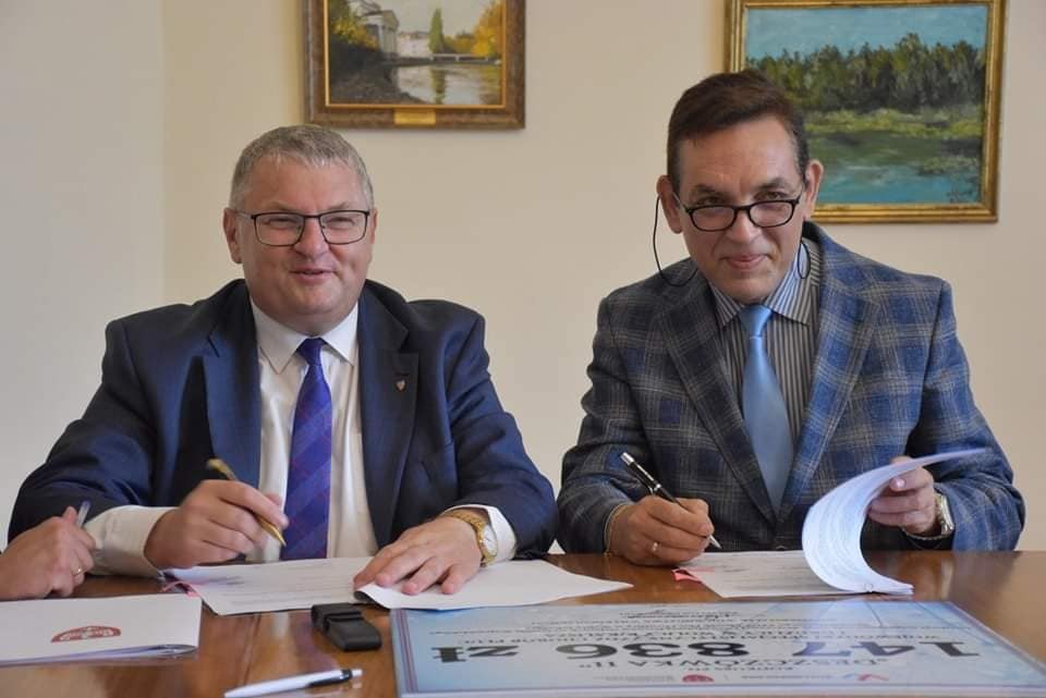 zdjęcie przedstawia mężczyzn podpisujących umowę. Po lewej Krzysztof Grabowski wicemarszałek województwa wielkopolskiego ubrany w niebieski garnitur, po prawej dyrektor szpitala w wolicy ubrany w marynakrę w niebieską kratę. na biurku rozłożone umowy, W rękach Panów długopisy
