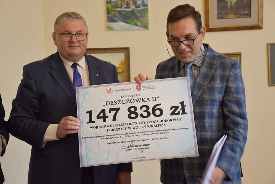 zdjęcie przedstawia dwóch mężczyzn wspólnie trzymających planszę informującą o przyznaniu dotacji w wysokości 147 836 złotych na realizację inwestycji w szpitalu. Po lewej Krzysztof Grabowski wicemarszałek województwa wielkopolskiego ubrany w niebieski garnitur, po prawej dyrektor szpitala w wolicy ubrany w marynarkę w niebieską kratę. 