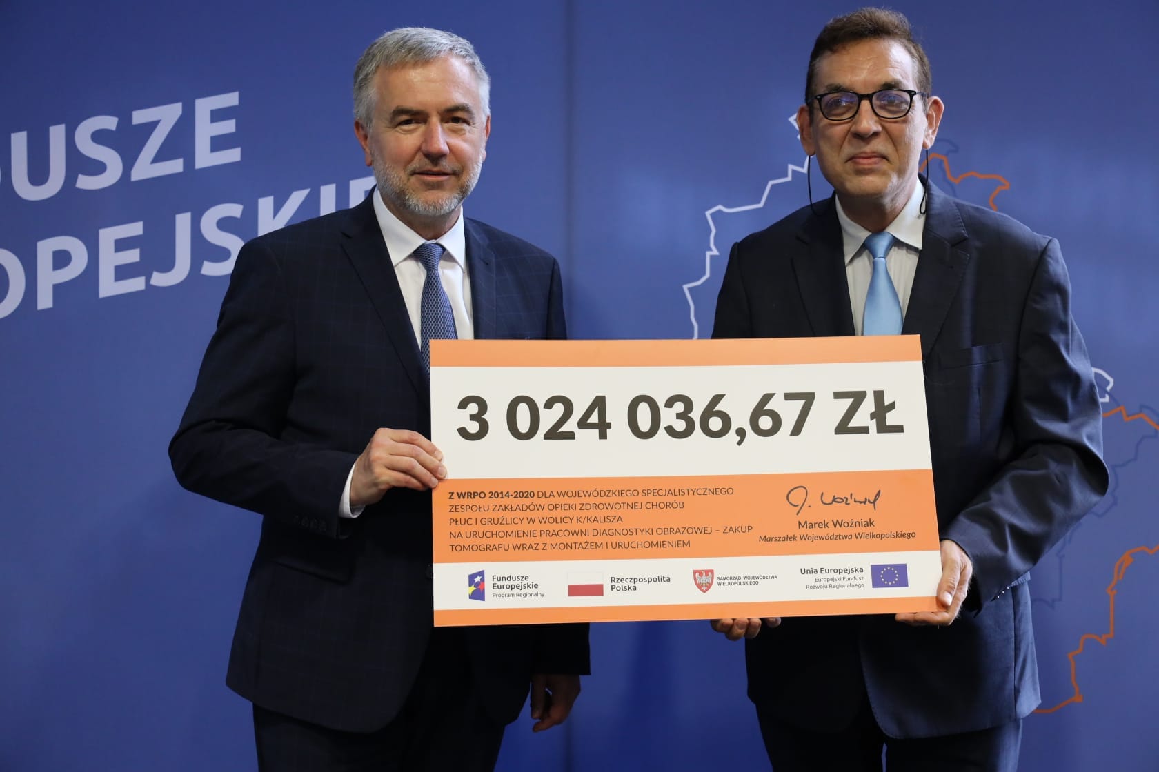 zdjęcie przedstawia Marszałka Województwa Wielkopolskiego Marka Wożniaka ubranego w czarny garnitur oraz Dyrektora szpitala w Wolicy Sławomira Wysockiego w granatowym garniturze trzymających wspólnie plansze z kwotą dofinansowania projektu wynoszącą 3024036,67 zł.